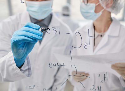 Close-up of two scientists writing chemical formulas on glass board and discussing them in team